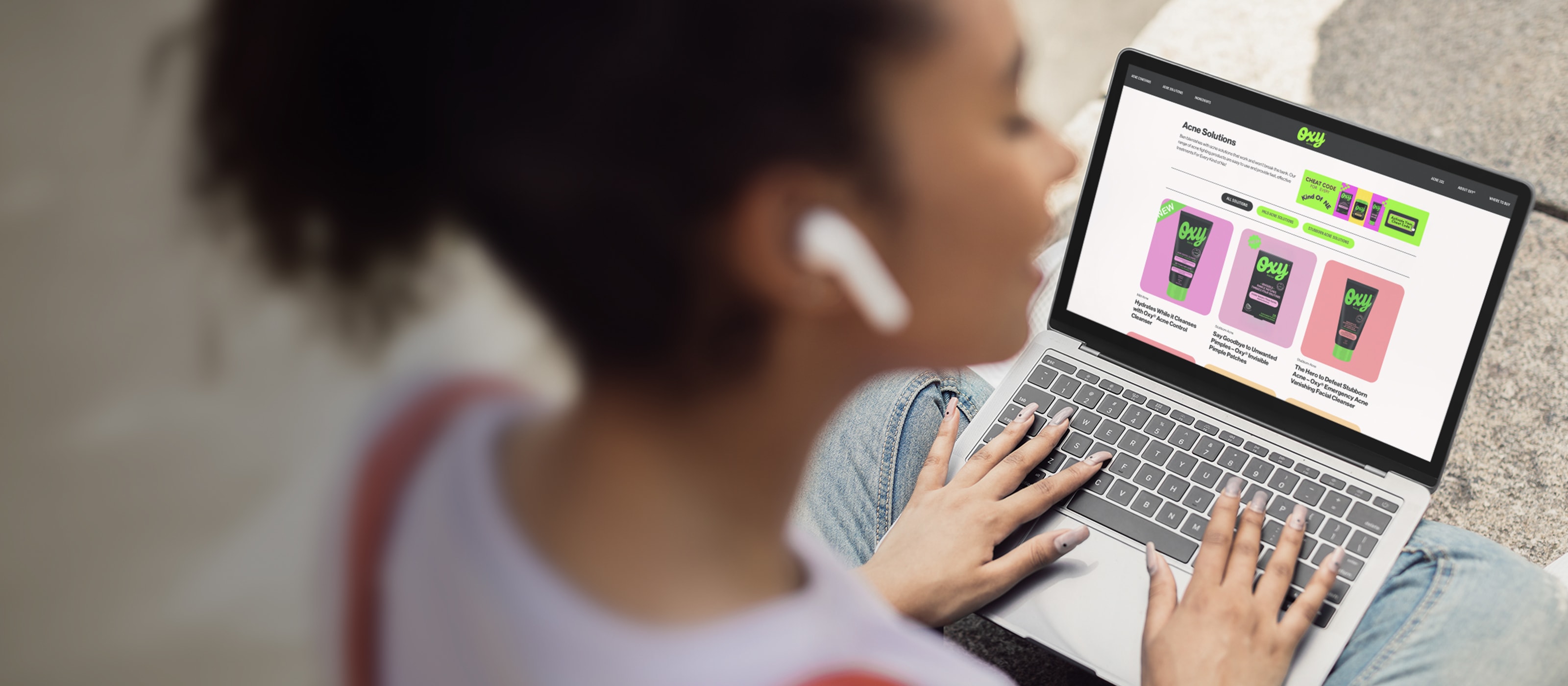 A girl wearing ear buds is on a laptop looking at the Oxy Acne Care website.